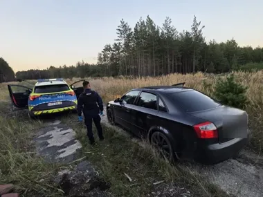 Policjant w niebieskich rękawiczkach podchodzi do czarnego sedanu stojącego na polnej drodze obok radiowozu policyjnego na tle lasu i łąki.