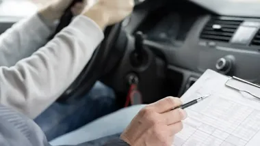 Zbliżenie na dłonie osoby trzymającej kierownicę samochodu oraz dłoń instruktora lub policjanta z długopisem i formularzem na podkładce.
